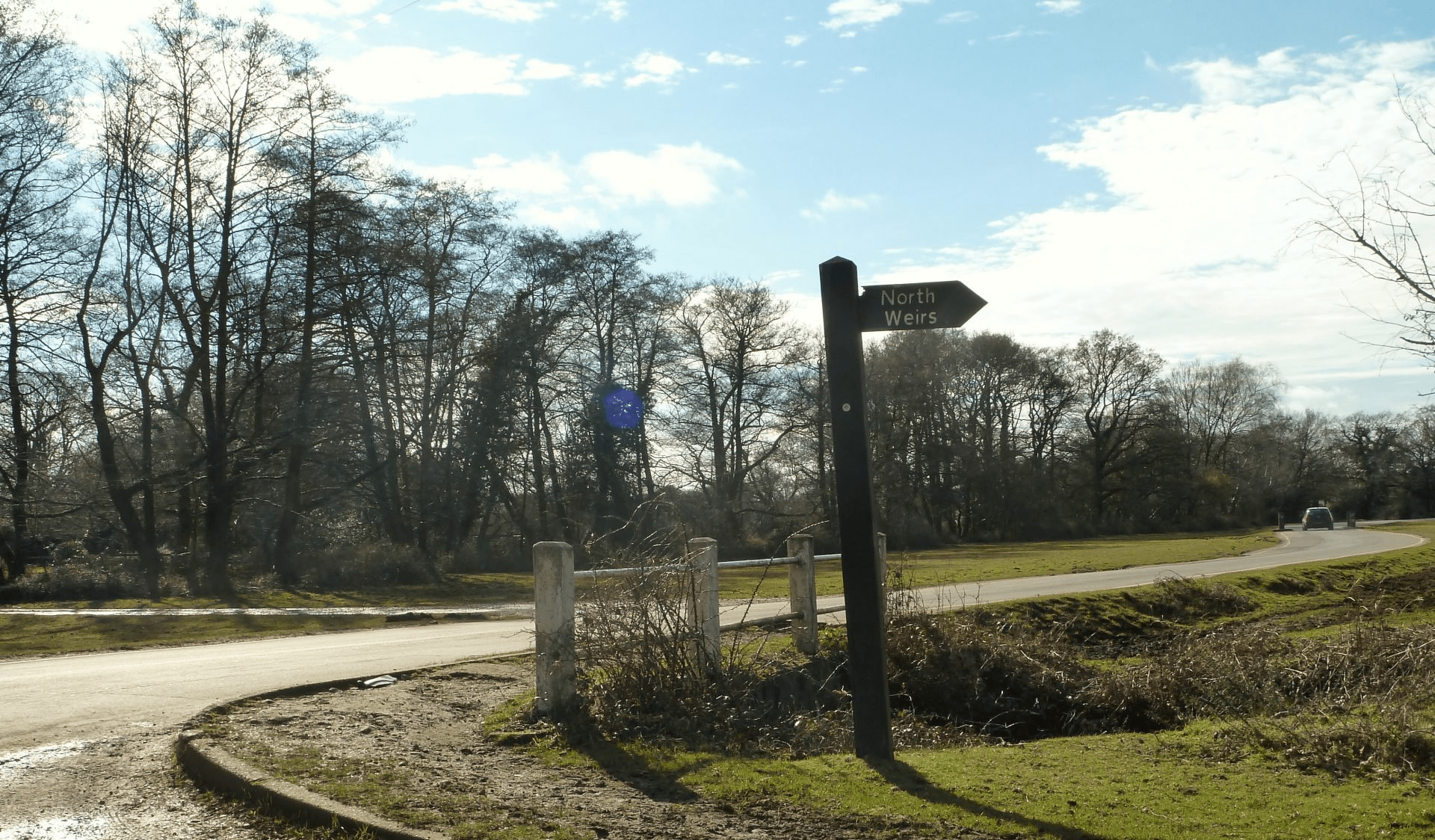 Roadside sign pointing to North Weirs beside a curving road, grassy verge, and leafless trees under a blue sky