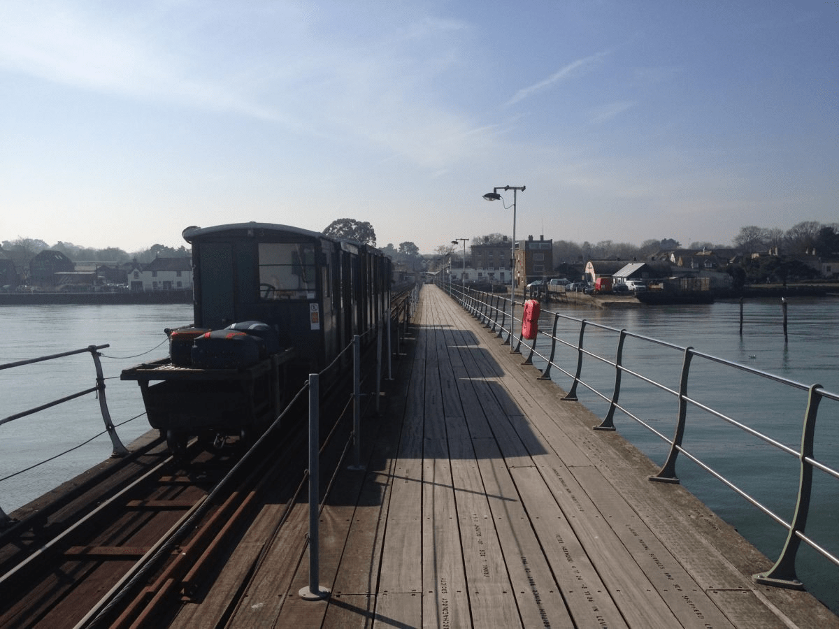 Train and wooden walkway on Hythe Pier over calm water, with railings, lifebuoy, and shoreline buildings in the distance
