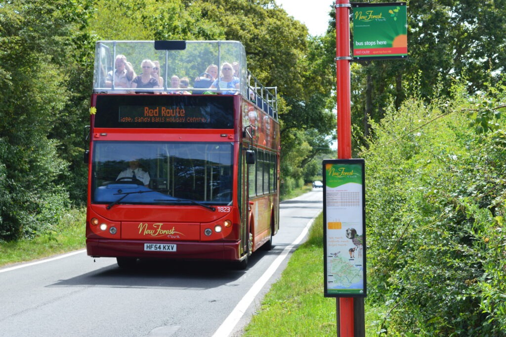Red open-top double-decker New Forest Tour bus on the Red Route passing a bus stop on a tree-lined country road