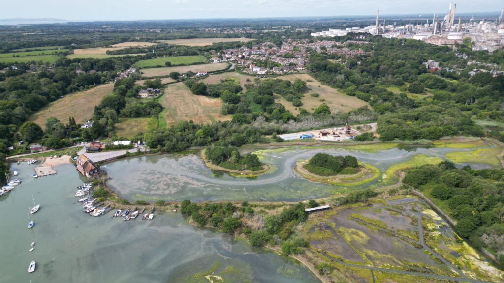 Aerial view of Fawley showing marshy river inlet with moored boats, wooded fields and housing, and an industrial refinery in the distance